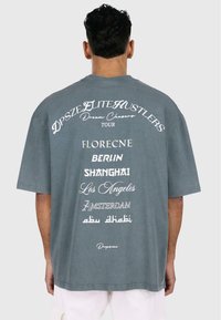 Grå oversized t-skjorte med hvit tekst på baksiden, som viser bynavn og "Dream Chasers Tour" i forskjellige skrifttyper.