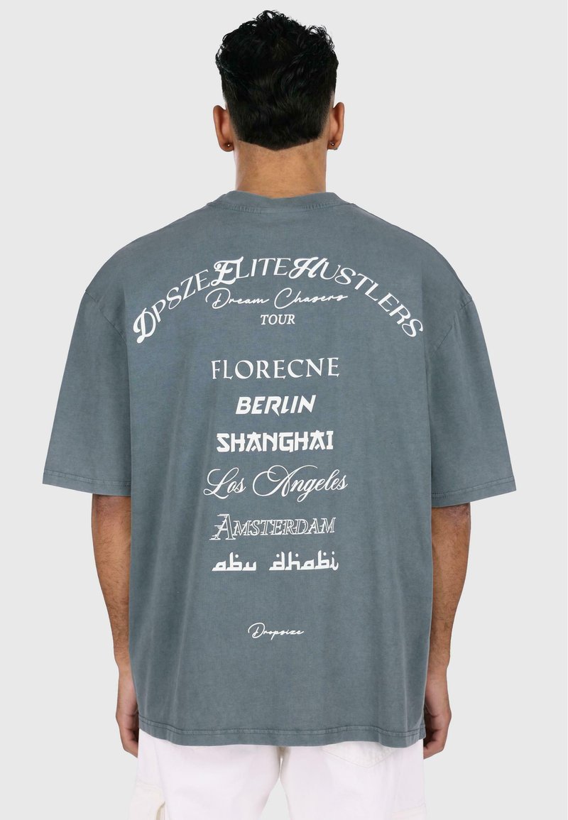 Sivá oversize tričko s bielym textom na chrbte, ktorý zobrazuje názvy miest a "Dream Chasers Tour" v rôznych písmach.