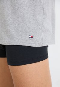 T-shirt en coton gris avec un petit logo rouge, blanc et bleu au bas, associé à un short noir ajusté, mettant en valeur une texture lisse.