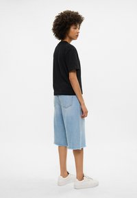 Juodasis medvilnės marškinėliai, laisvas pritaikymas, derinami su šviesiai mėlynais džinsiniais šortais, suplėšytu kraštu, mūvimi baltais sportbačiais. Modelis stovintis šonu.