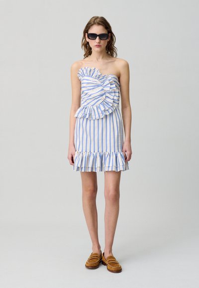 Abito senza spalline con righe verticali blu e bianche, dettagli arricciati e orlo a frange. Indossato con occhiali da sole e mocassini marroni.