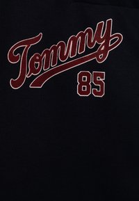 T-shirt in cotone nero con un importante logo rosso in corsivo "Tommy" e il numero "85" in rosso, su una superficie testurizzata.