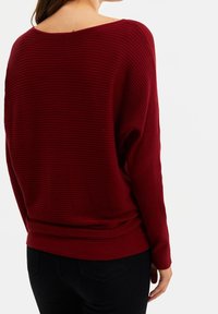 Pull en tricot côtelé bordeaux avec une coupe décontractée, un large col et des manches longues. Texture douce avec un ourlet loose. Porté avec un pantalon noir.