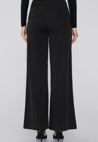 Persona con pantalones vaqueros negros de cintura alta y pierna ancha y sandalias de tacón negro, de pie con los brazos relajados a los lados.