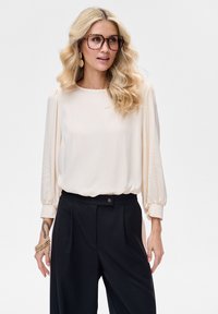 Jasnobeżowa bluzka z długimi bufiastymi rękawami, gładką teksturą i okrągłym dekoltem, zestawiona z czarnymi spodniami z wysokim stanem, zapinanymi na guziki.