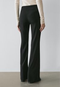 Woman wearing black high-waist wide-leg pants and a beige long-sleeve top, standing on a gray floor against a plain gray wall.