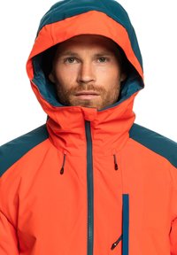 Veste imperméable orange vif avec capuche bleue et accents d'épaule. Elle possède un col montant, des cordons de serrage réglables et une fermeture éclair à l'avant.
