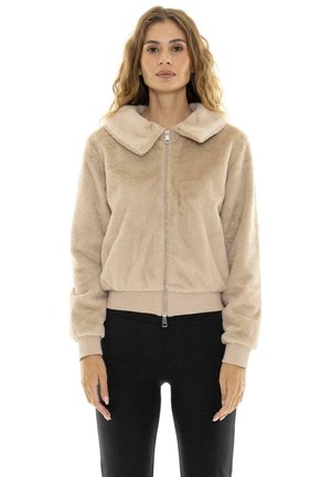 Une femme présente une veste beige en peluche, zippée et à col large, avec un pantalon noir, sur fond blanc.