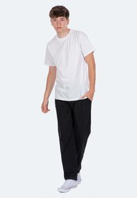 Weißes Baumwoll-T-Shirt, kombiniert mit schwarzen, lockeren Hosen und weißen Sneakers. Das Modell steht vor einem neutralen Hintergrund.