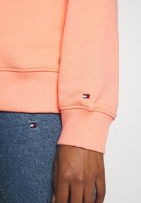Lichtroze sweatshirt met ribgebreide manchetten, gecombineerd met denim voorzien van kleine geborduurde logoaccenten in rood, wit en blauw.