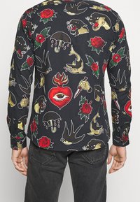 Chemise noire à manches longues avec des imprimés colorés inspirés des tatouages, comprenant des roses, des panthères et des motifs nautiques en rouge, jaune et vert.
