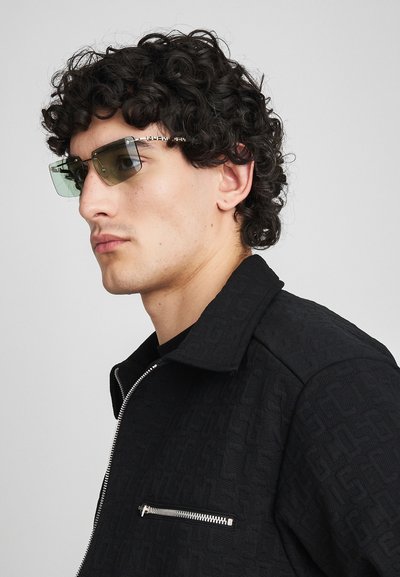 Chaqueta negra con estampado y un bolsillo con cremallera, combinada con gafas de sol angulares de tintado verde con acentos metálicos.