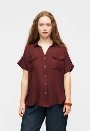 Νεαρή γυναίκα με μακριά κόκκινα μαλλιά, φορώντας μια κουμπωτή μακρυμάνικη μπλούζα μπορντό χρώματος και σκούρα μπλε παντελόνια, στέκεται μπροστά σε ένα απλό φόντο.