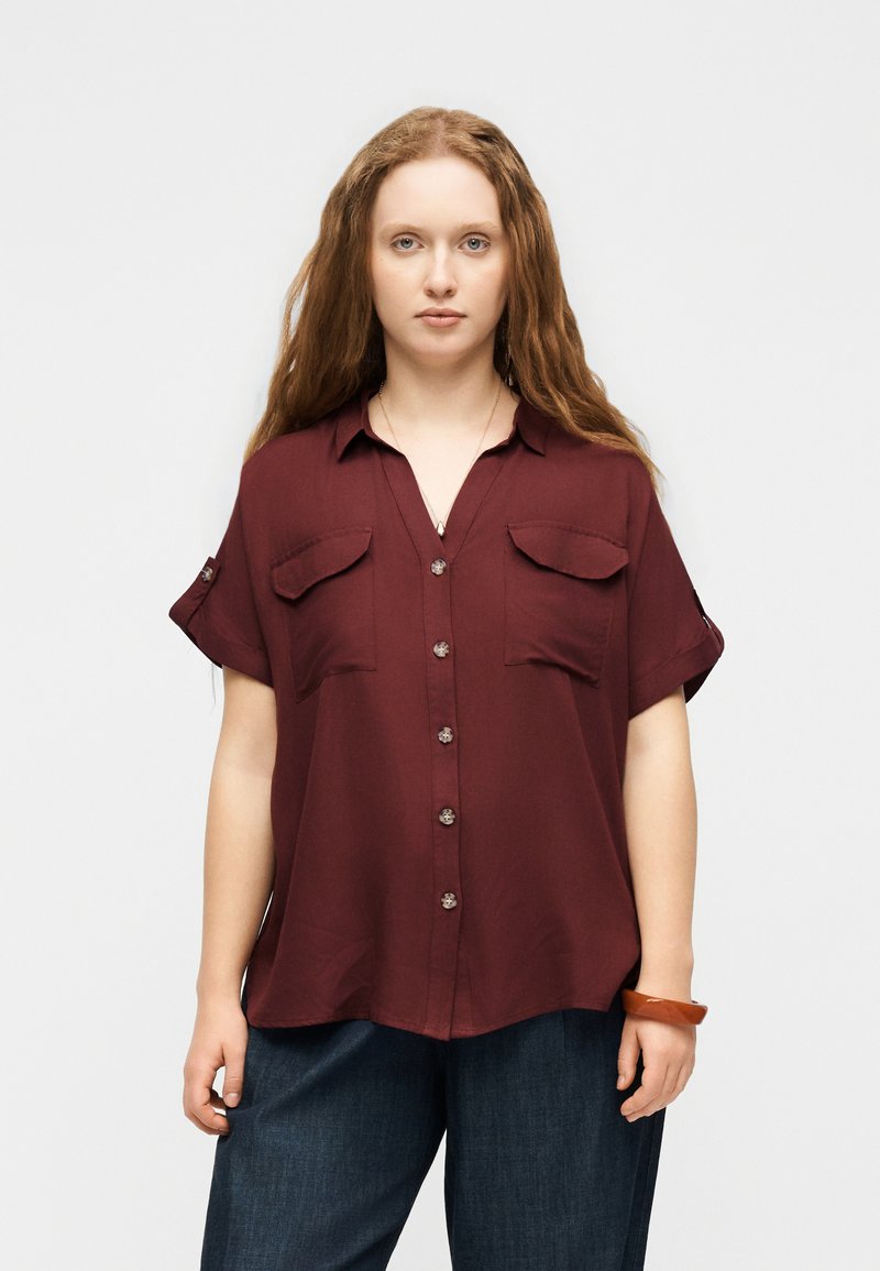 Νεαρή γυναίκα με μακριά κόκκινα μαλλιά, φορώντας μια κουμπωτή μακρυμάνικη μπλούζα μπορντό χρώματος και σκούρα μπλε παντελόνια, στέκεται μπροστά σε ένα απλό φόντο.