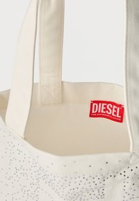 Τσάντα ώμου από καμβά σε εκρού χρώμα με ασημένια διακοσμητικά καρφάκια. Διαθέτει ανθεκτικό ύφασμα, δύο παράλληλες λαβές και κόκκινο λογότυπο Diesel στο εσωτερικό.