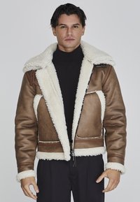 Chaqueta de imitación de piel de oveja marrón con un cuello de forro polar blanco, cremallera expuesta y acentos en contraste. Presenta bolsillos laterales y una silueta ajustada.