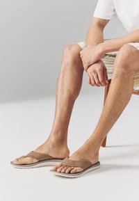 Braune Flip-Flops mit einem Obermaterial aus strukturiertem Stoff und einer gepolsterten Sohle, mit minimalistischer Gestaltung und offenem Zehenbereich.