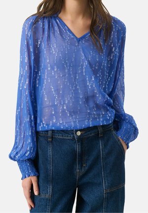 Femme portant un chemisier bleu transparent à taches argentées avec des poignets smockés, rentré dans un jean denim bleu foncé taille haute.