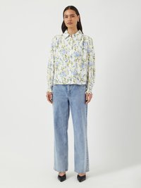 Blommig knäppblus med blå och gula blommor på vit bakgrund, veckade ärmar, bärs med ljusblå vida jeans och svarta klackar.