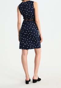 Dunkelblaumeliertes Kleid mit tailliertem Schnitt, ärmellosem Design und einem Gürtel, aus glattem Stoff und bis zur Mitte des Oberschenkels reichend, kombiniert mit schwarzen Schuhen.