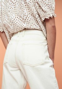 Jeans in denim bianco con vita alta, caratterizzati da tasche posteriori e cuciture a contrasto, abbinati a una blusa ricamata con texture.