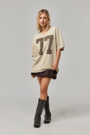 T-shirt beige oversize avec un graphisme "77" marron foncé. Le modèle porte une jupe noire à volants et des bottes noires arrivant au genou. Texture de tissu lisse.