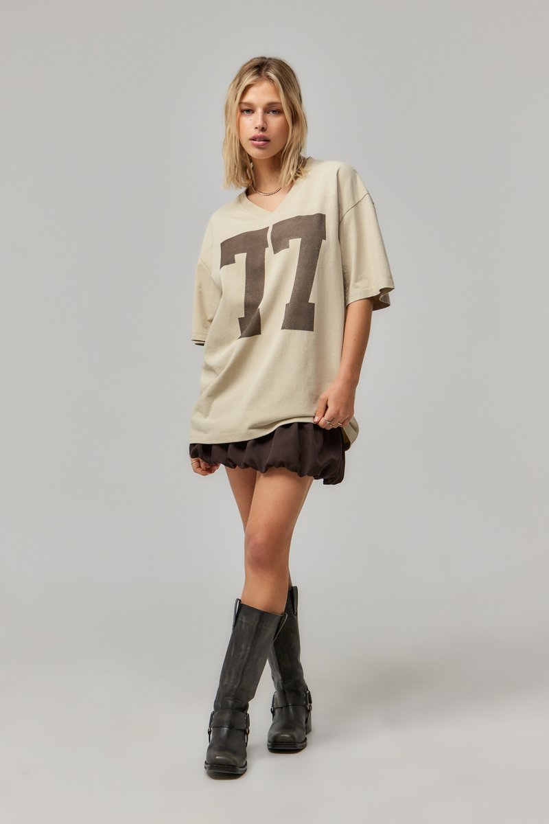 Beige oversized t-shirt met een donkerbruine "77" graphics. Model draagt een zwarte geruite rok en hoge zwarte laarzen. Soepele stofstructuur.