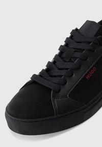 Black sneaker with textured side panels, thick black laces, and red "HUGO" logo on the side, shown against a white background.
