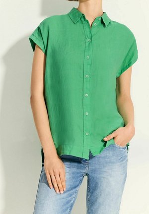 Femme portant une chemise verte à manches courtes avec col et boutons, un jean bleu clair, une main dans la poche, debout devant un fond clair uni.