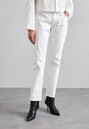 Person iført hvide straight-leg jeans og sorte ankelstøvler med spids tå, stående på en grå gulv mod en lys væg.