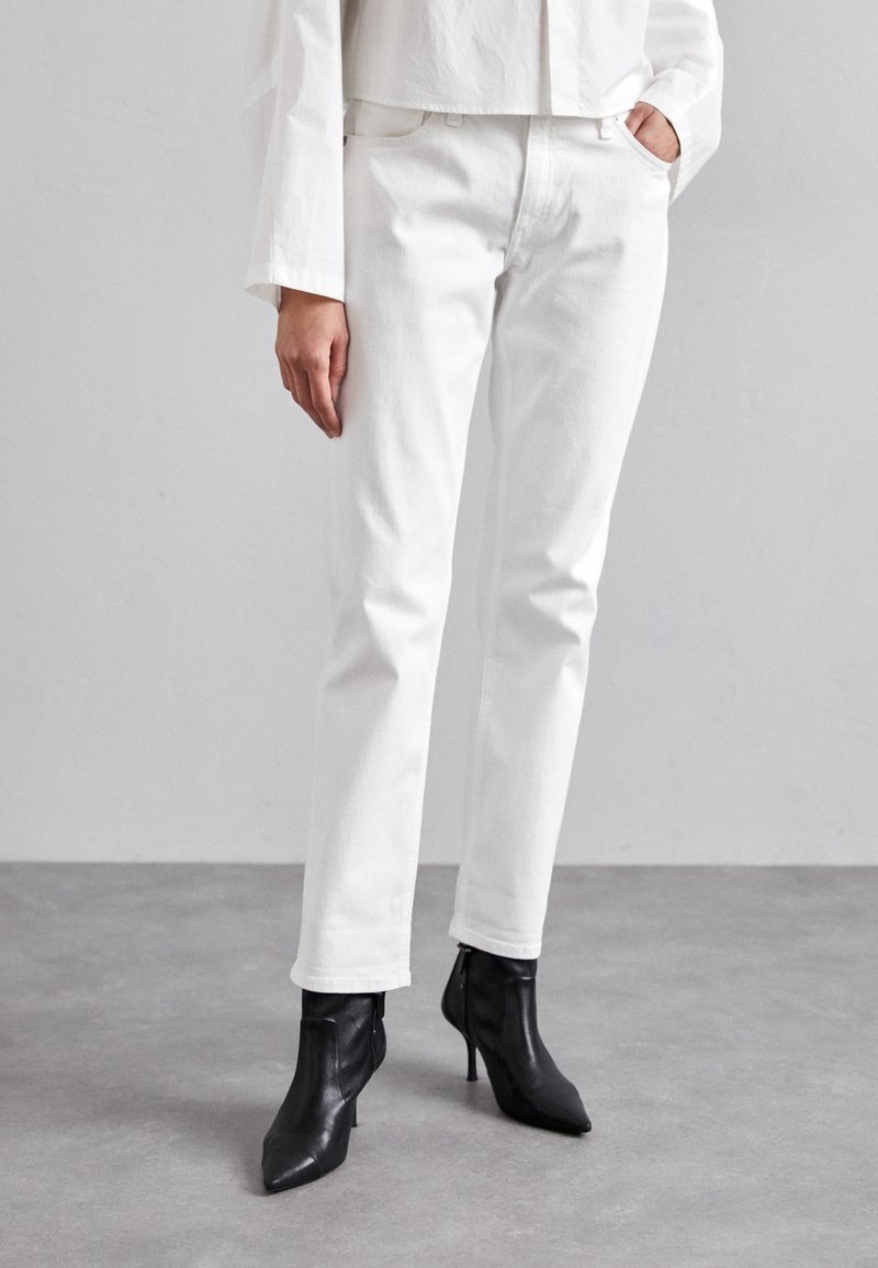 Personne portant un jean blanc droit et des bottines noires à bout pointu, debout sur un sol gris contre un mur clair.
