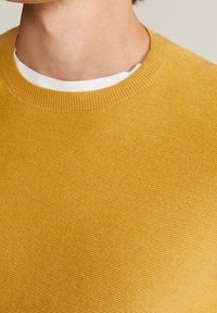 Hořčicově žlutý pletený svetr, kulatý žebrovaný výstřih, texturovaná tkanina, detailní pohled na vzor pletení a límec.