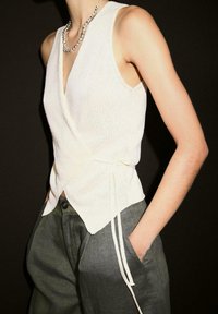 Maglione avvolgente leggermente beige, lavorato a maglia, con scollatura a V e lacci laterali, indossato sopra pantaloni larghi in lino grigio scuro, caratterizzati da una leggera trama e gamba ampia.