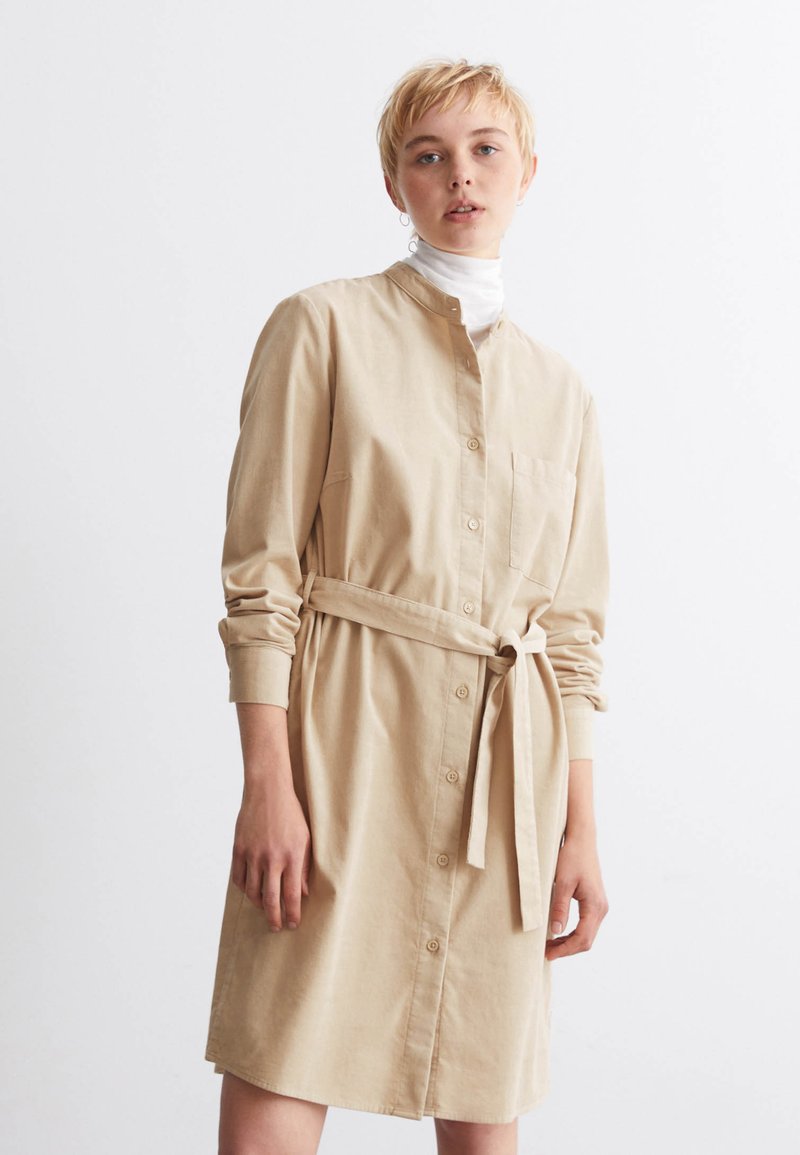 Beige Hemdkleid mit langen Ärmeln aus einem weichen Stoff. Es hat einen Kragen, vordere Knöpfe, eine einzelne Brusttasche und einen gebundenen Taillengürtel.