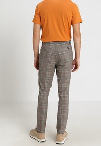 Pantalones de cuadros grises con un corte ajustado, con acentos rojos y un frente plano, hechos de una tela texturizada. Llevados con una camisa de manga corta naranja.