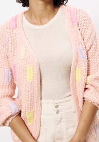 Světle růžový pletený kardigan s vícebarevnými listovými motivy, vrstvený přes béžové žebrované tričko, zkombinovaný s krémovými vysokým pasem kalhot.