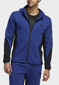 Veste de sport zippée bleu avec accents noirs, présentant un tissu texturé, une coupe ajustée et un logo brodé sur la poitrine.