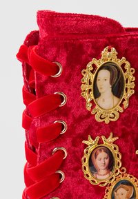 Botte en velours rouge avec une surface texturée, ornée de portraits encadrés en or sur le côté et de lacets en satin rouge décoratifs.
