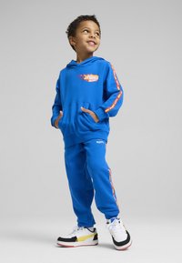 Niebieska bluza z kapturem z kieszenią z przodu i płomieniami, zestawiona z dopasowanymi niebieskimi spodniami dresowymi. Posiada kolorowe logo i białe sneakersy z akcentami.