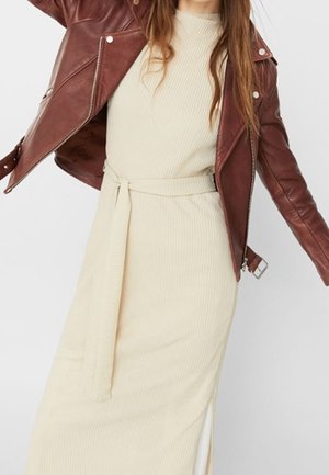 Mujer con vestido de punto acanalado color crema con cinturón en la cintura y chaqueta de cuero marrón con detalles de cremallera y hebilla.