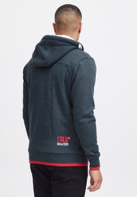 Sweat-shirt en tissu gris foncé à finition texturée, doté d'une capuche, d'un accent rouge à l'ourlet et d'un logo au dos près de la taille.