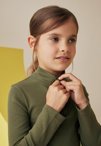 Długi rękaw, oliwkowa zielona bluza na zamek z prążkowaną fakturą. Wysoki kołnierz i dopasowany krój. Dobrze komponuje się z warstwowymi strojami lub casualowym ubiorem.