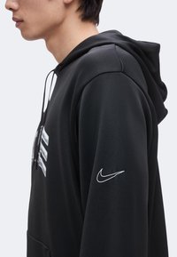 Jeune personne portant un sweat à capuche noir avec le logo Nike blanc sur la manche, vue de profil sur un fond uni.