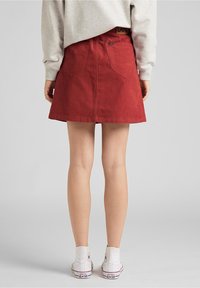 Falda de pana roja con un dobladillo acampanado, que presenta bolsillos con parche y una etiqueta con el logotipo en la cinturilla. Combinada con zapatillas de lona blancas.