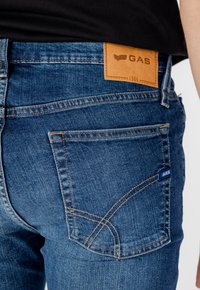Jeans in denim blu con texture liscia, cuciture a contrasto e un'etichetta in cuoio marrone sulla vita con la scritta "GAS" e l'anno "1984."