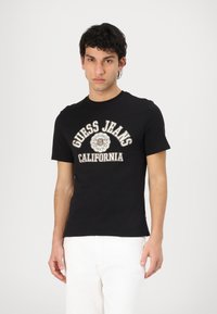 Camiseta de algodón negra de manga corta con el logo "Guess Jeans California" en blanco y marrón. Corte ajustado con escote redondo.