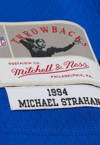 Patch rétro avec un graphisme de joueur de football, étiquette Mitchell & Ness Nostalgia Co., et "1994 Michael Strahan" cousu sur un tissu bleu.