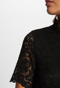 Zwarte kant top met een hoge halslijn, korte mouwen, bloemmotieven en gegolfde randen, die ingewikkelde texturen en transparantie laat zien.