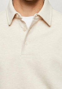 Camisa polo de tela crema con una textura suave. Presenta un cuello clásico, tapeta de tres botones y un sutil detalle de costura en el cuello.
