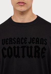 Schwarzes Baumwoll-T-Shirt mit fetter, erhabener schwarzer Schrift "VERSACE JEANS COUTURE". Klassisches Rundhalsdesign mit glatter Textur.
